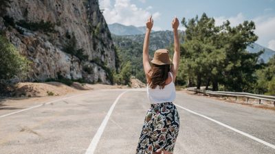 A woman with arms raised stands joyfully on a mountain road in summer.